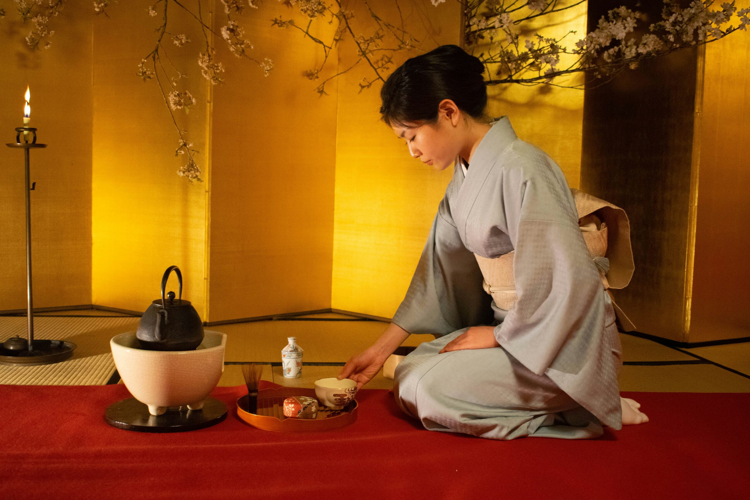 A Japanese tea ceremony with a woman in kimono kneeling beside a traditional tea set, framed by cherry blossoms and candlelight.
